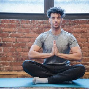 Full body of focused male sitting in Sukhasana with praying hands while practicing yoga on sports mat
